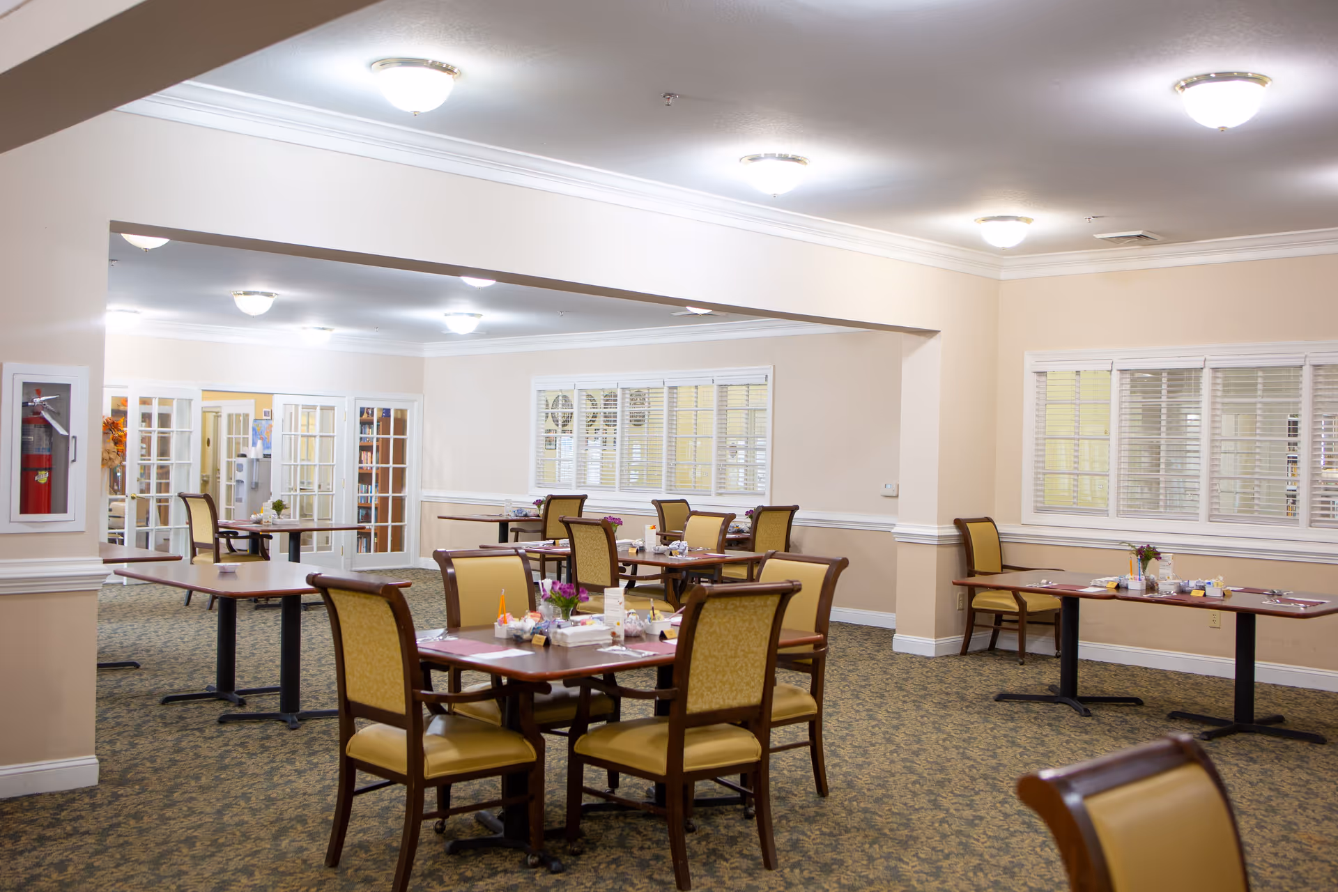 A spacious dining room with multiple wooden tables and cushioned chairs arranged neatly. The room has beige walls, carpeted floor, and several ceiling lights. There are windows with white blinds and glass doors leading to another room. Tables are set with placemats, napkins, and small flower arrangements.