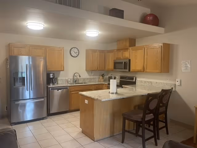 Open kitchen with stainless steel refrigerator and dishwasher, wooden cabinets, a center island with two chairs, and tiled floor.