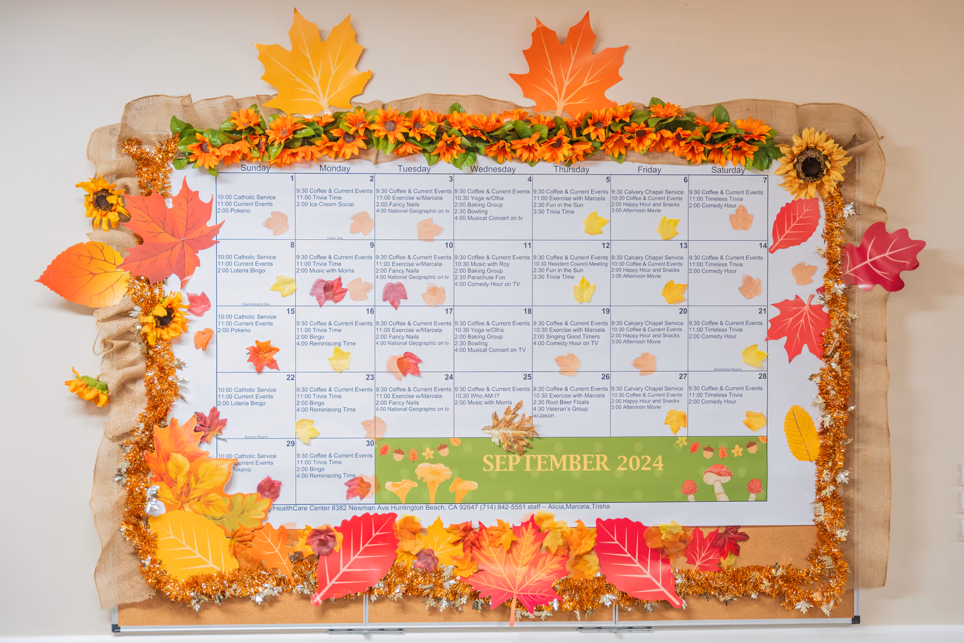A bulletin board decorated with autumn-themed leaves, flowers, and garlands surrounding a calendar for September 2024. The calendar lists daily activities and events at Huntington Valley Healthcare, including services like Catholic Service, Bingo, Coffee & Current Events, Exercise, and Movie times.