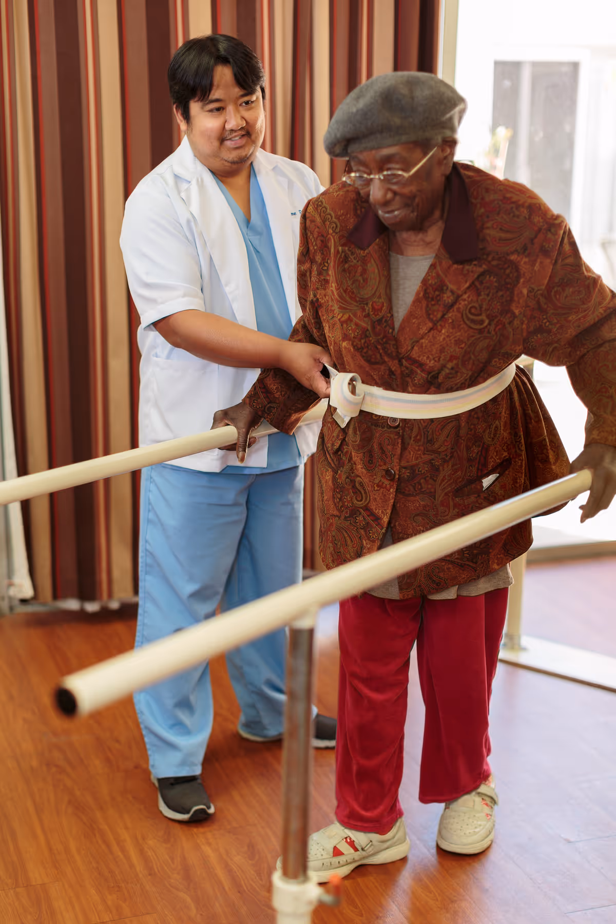 A healthcare worker in blue scrubs and a white coat assists an elderly woman wearing a patterned jacket, red pants, and a gray beret as she practices walking between parallel bars in a rehabilitation or therapy setting.