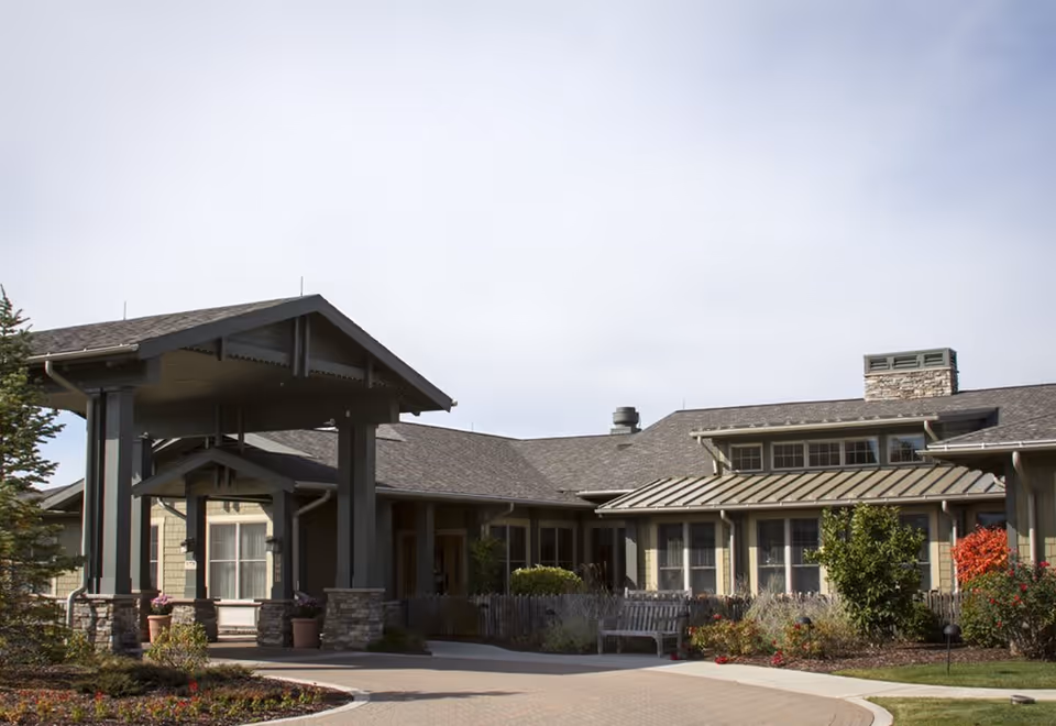 Exterior view of a single-story senior living facility building with a covered entrance supported by stone pillars. The building has a mix of stone and siding exterior, multiple windows, and a landscaped garden with bushes and flowers in front.