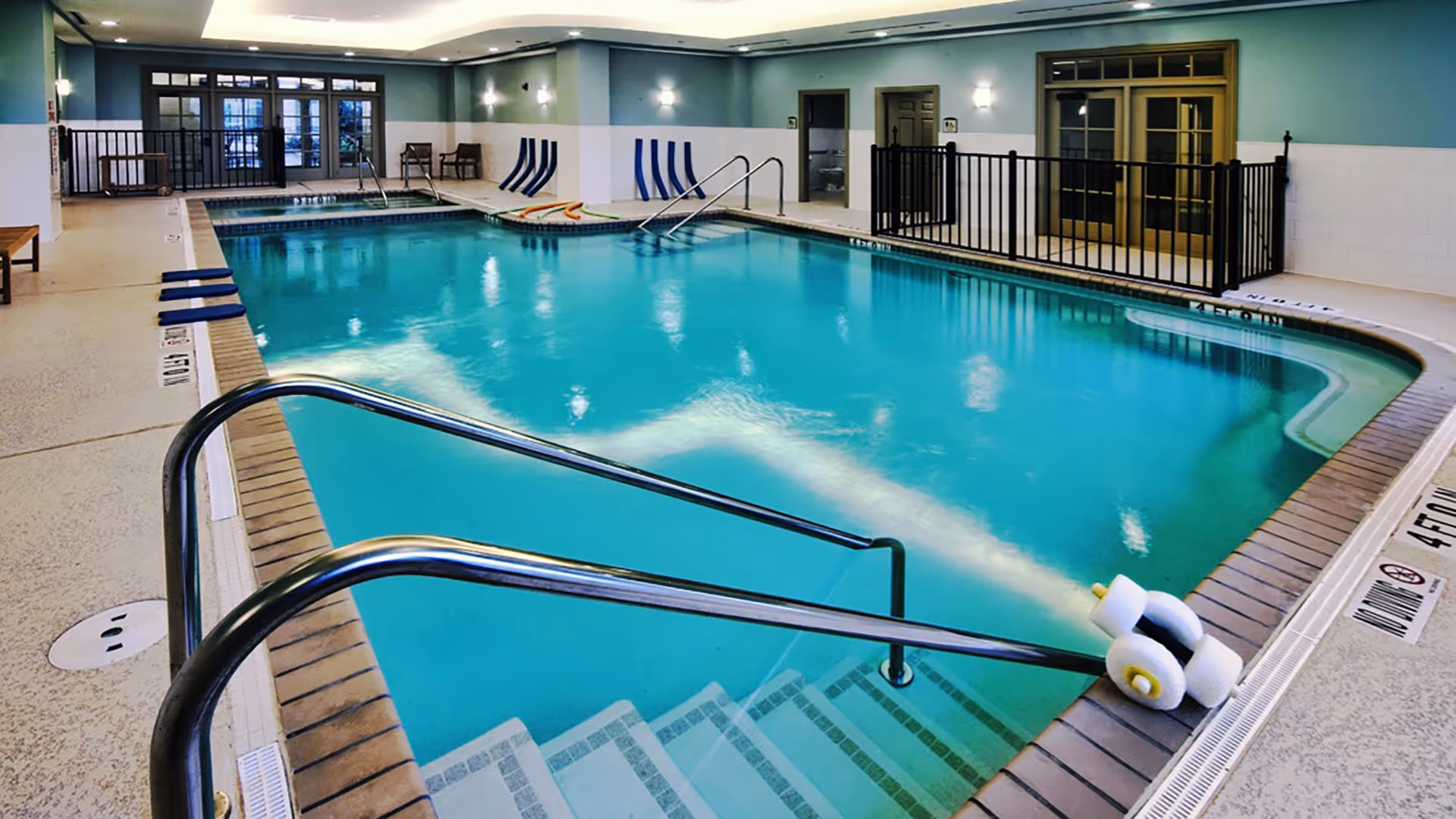 Well-lit indoor swimming pool with metal handrails, entry steps, lounge chairs and a fenced doorway area.