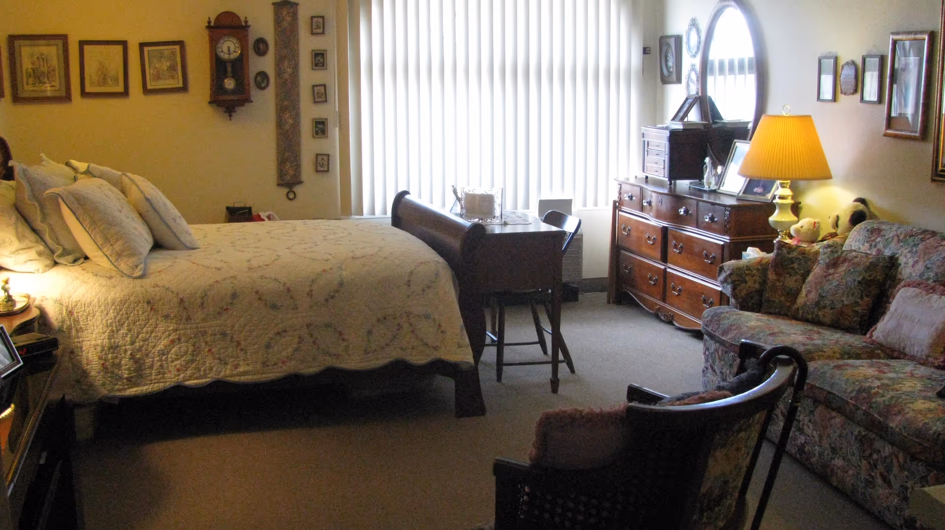 Sunlit bedroom with a bed, wooden dresser, small desk, and a floral sofa seating area.