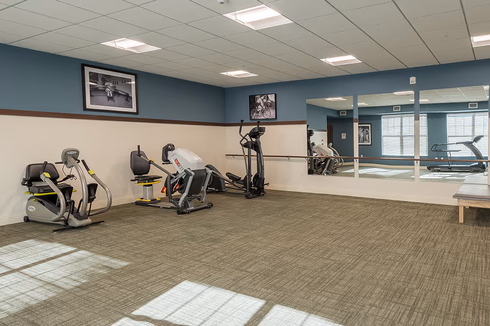 A fitness room with exercise equipment including a recumbent bike, an elliptical machine, and other workout machines. The room has blue and beige walls, a large mirror on one side, framed black and white photos on the walls, and carpeted flooring with sunlight streaming through windows.