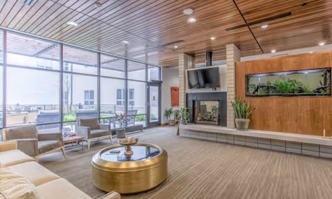 A spacious and modern living room area with large floor-to-ceiling windows allowing natural light to fill the space. The room features a beige sofa, several armchairs, a round gold coffee table, a fireplace with a mounted TV above it, and a built-in aquarium on the wooden wall. There are plants placed around the room and an outdoor patio visible through the windows.