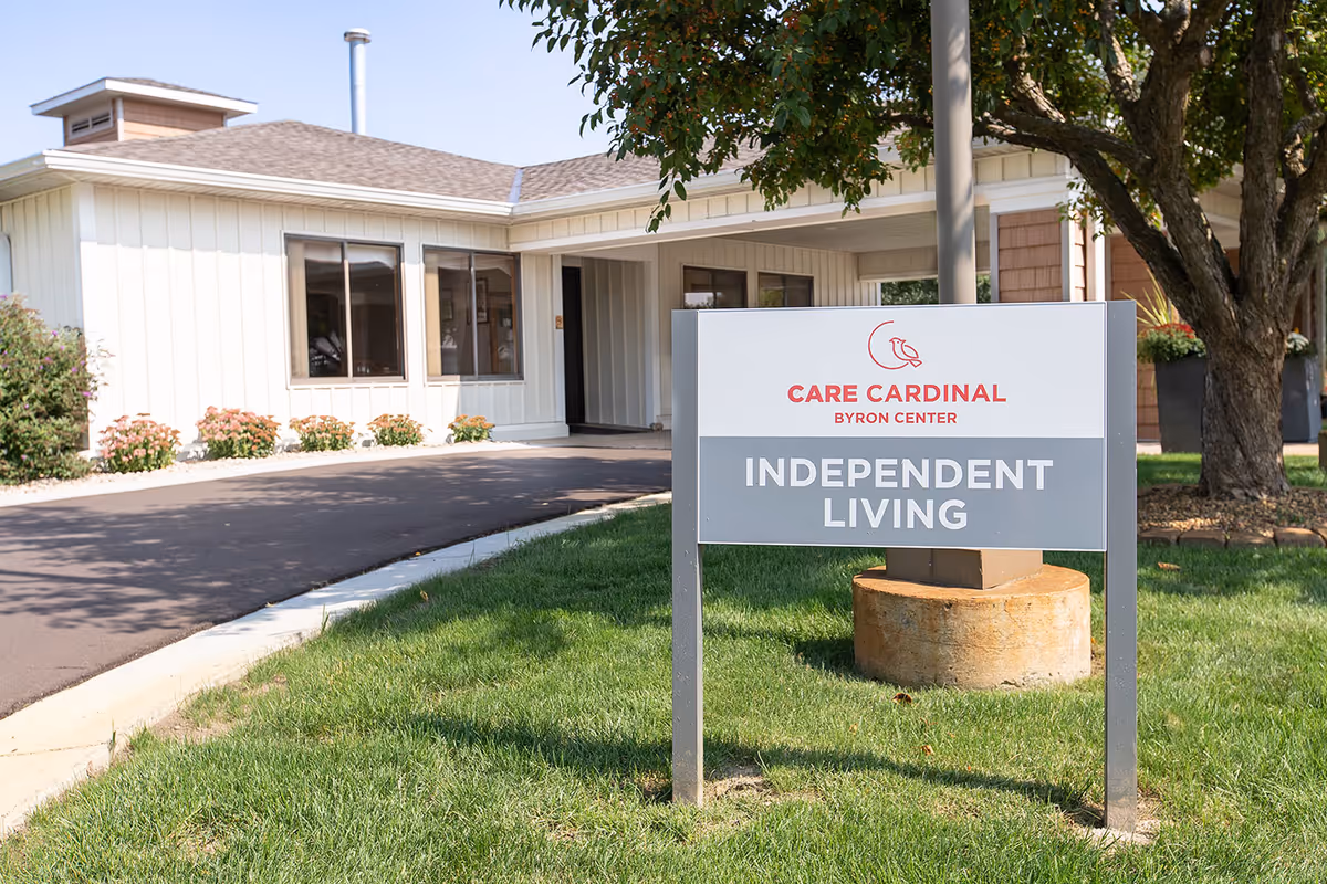 Exterior view of Care Cardinal Byron Center building with a sign in the foreground that reads 'Care Cardinal Byron Center Independent Living'. The building has light-colored siding, several windows, and a driveway leading to the entrance. There is a tree and green grass around the sign.