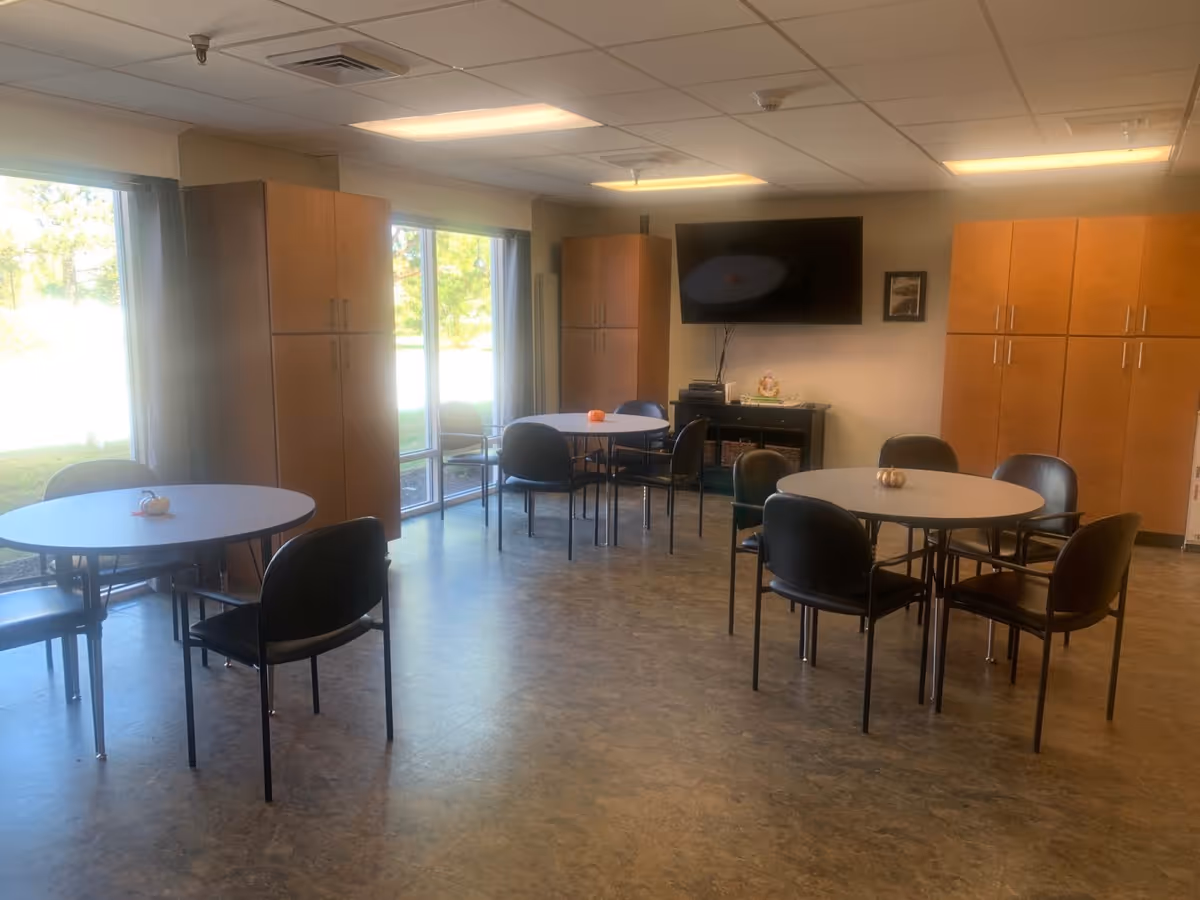 A well-lit room with round tables and black chairs arranged around them. Each table has a small decorative pumpkin. Large windows on one side let in natural light and show greenery outside. There are tall wooden cabinets against the walls and a flat-screen TV mounted on the wall above a small black cabinet.