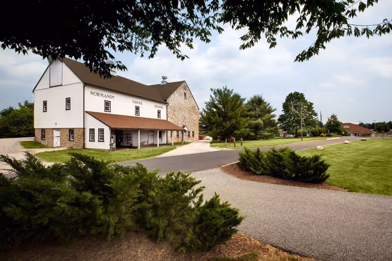 A large white and stone building with a brown roof labeled Normandy Farms Estates, surrounded by green bushes, trees, and a paved driveway under a partly cloudy sky.