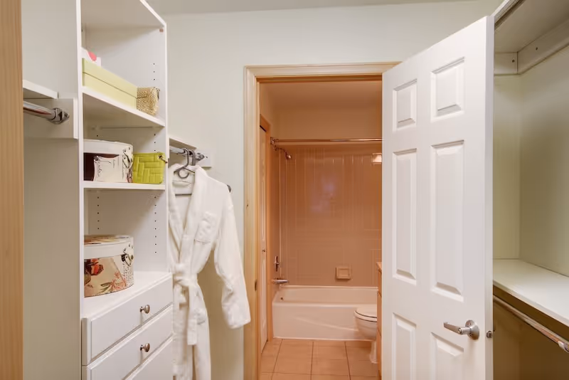 View of a walk-in closet with white shelves and hanging rods, featuring a white bathrobe hanging on a hook. Through an open door, a bathroom is visible with a bathtub, shower curtain rod, and toilet.