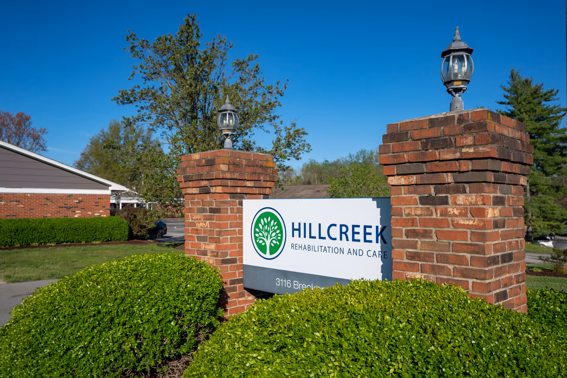 Outdoor view of the entrance sign for Hillcreek Rehabilitation and Care, mounted between two brick pillars with lanterns on top, surrounded by green bushes and trees under a clear blue sky.