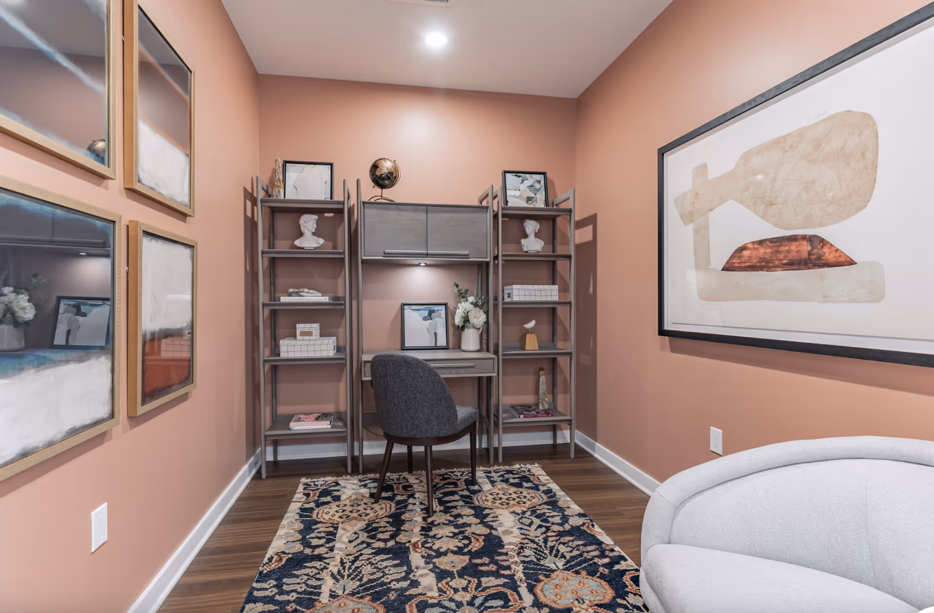 A small study or office area with peach-colored walls, featuring a gray desk with shelving units on either side. The desk has decorative items including bust sculptures, framed pictures, and a globe. A dark gray upholstered chair is placed in front of the desk on a patterned rug. The room also has framed abstract art on the walls and a light gray curved sofa partially visible on the right.