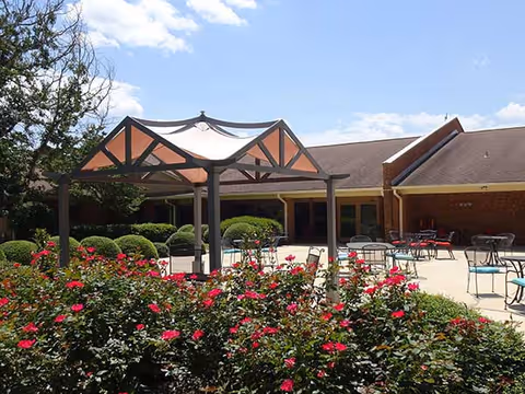 Outdoor courtyard with a central fabric canopy, patio tables and chairs, and pink flowering shrubs in front of a brick building.