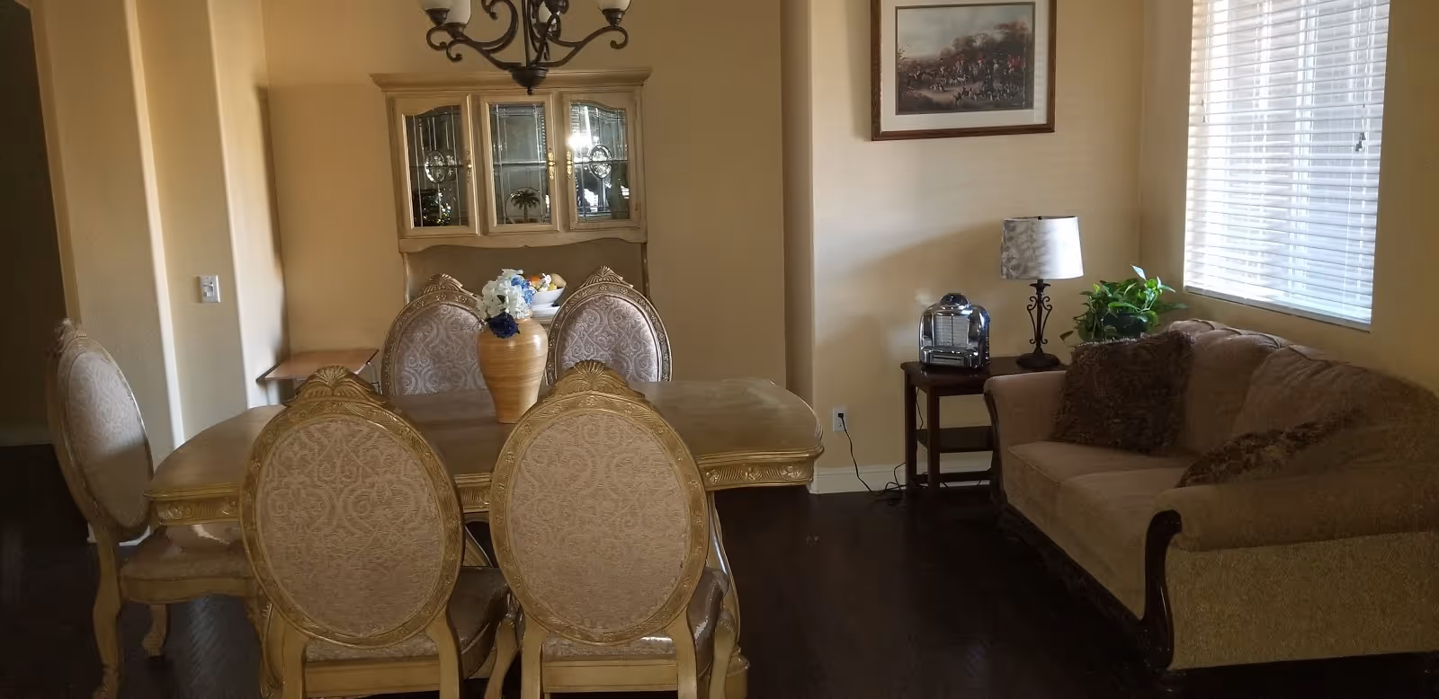 A cozy interior room featuring a dining area with a wooden table and six upholstered chairs. A decorative vase with flowers is placed on the table. Behind the table is a glass-fronted cabinet. To the right, there is a beige sofa with cushions, a side table with a lamp and a small plant, and a window with blinds allowing natural light to enter. A framed picture hangs on the wall above the side table.