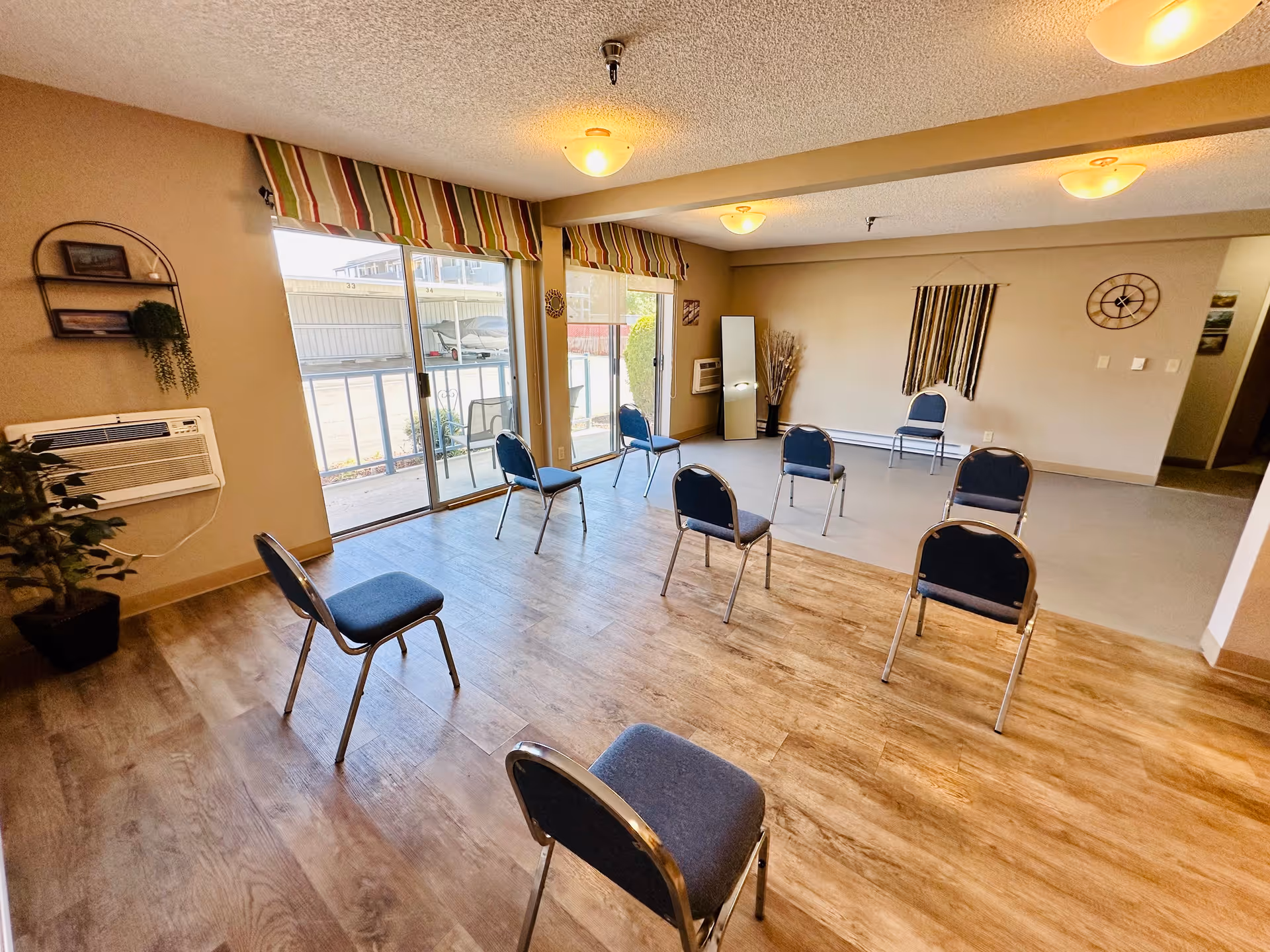 A spacious room with wooden and tiled flooring featuring eight blue chairs arranged in a circle. The room has beige walls, three ceiling lights, a wall clock, a tall mirror, and two large sliding glass doors with striped valances that let in natural light. There are some decorative plants and wall hangings, and an air conditioning unit is mounted on the left wall.