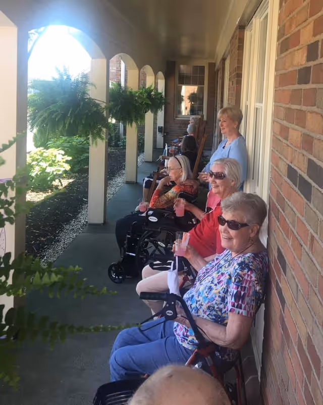 A group of elderly women sitting on chairs and wheelchairs along a covered outdoor walkway with brick walls and hanging plants, enjoying drinks and smiling on a sunny day.