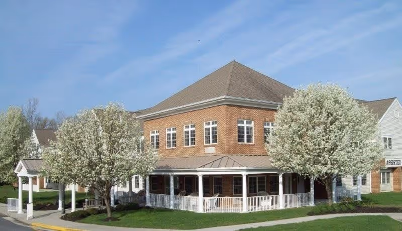 A two-story brick building with a wraparound porch surrounded by green grass and blooming white-flowered trees under a clear blue sky.