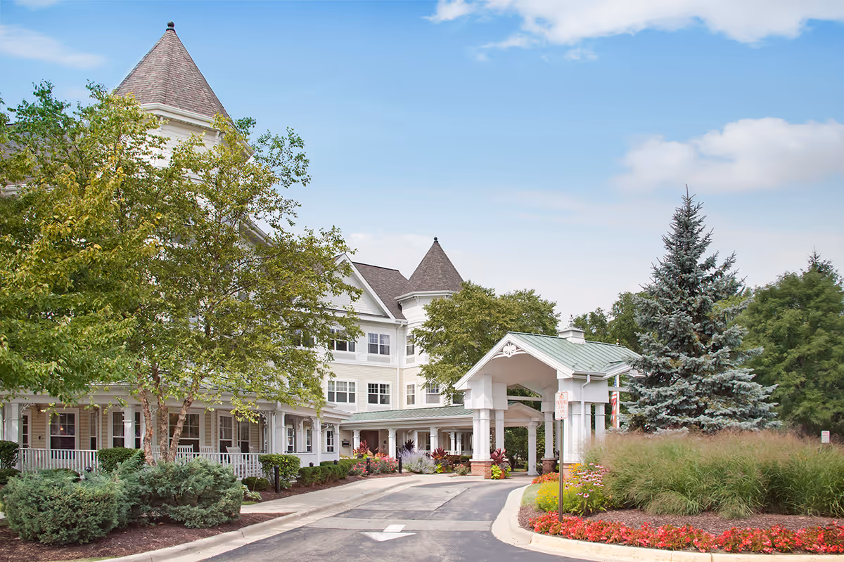 Front entrance of a large white multi-story senior living building with a porte-cochere, turreted roof, driveway, and landscaped grounds.