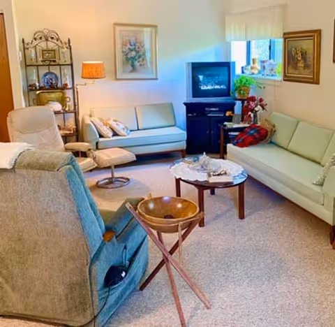 A cozy living room with two light-colored sofas, a beige recliner chair, a small round wooden coffee table with decorative items, a wooden side table with a lamp, a TV on a stand, and framed artwork on the walls. The room has a carpeted floor and a window with a white curtain.