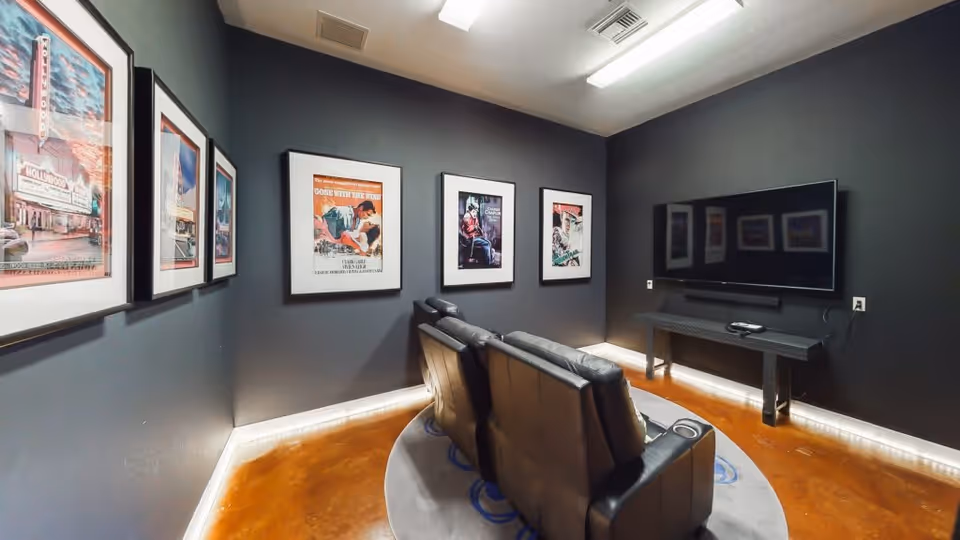 A small home theater room with three black leather recliner chairs arranged in front of a wall-mounted flat-screen TV. The walls are painted dark gray and decorated with framed vintage movie posters. The floor is polished wood with a circular rug beneath the chairs, and there is subtle LED lighting along the base of the walls.