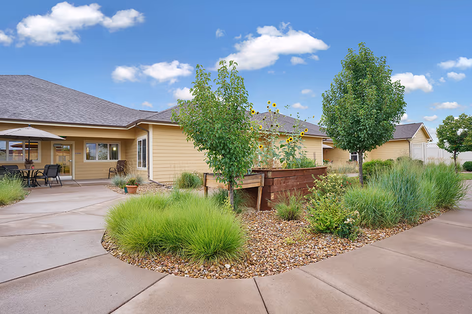 Outdoor view of a senior living facility with a paved walkway, green shrubs, small trees, and a raised garden bed with sunflowers. The building has beige siding and a gray roof under a blue sky with scattered clouds. There is a patio area with a table, chairs, and an umbrella.