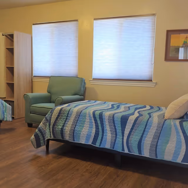 A simple bedroom with a single bed covered in a blue patterned quilt, a green armchair, two windows with blinds, and wood flooring.