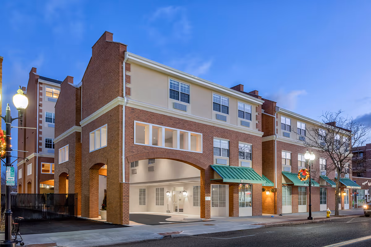 Exterior view of a multi-story senior living facility building at dusk with brick and beige walls, multiple windows, green awnings, street lamps decorated with holiday wreaths, and a driveway entrance under an archway.