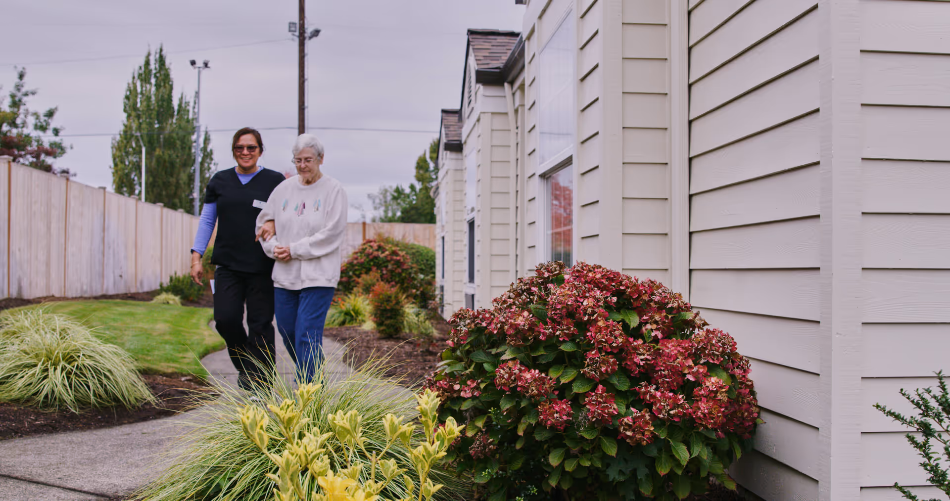 An elderly woman is walking outside along a garden path, supported by a caregiver. They are next to a beige building with large windows and surrounded by green plants and bushes with red flowers. The sky is overcast.