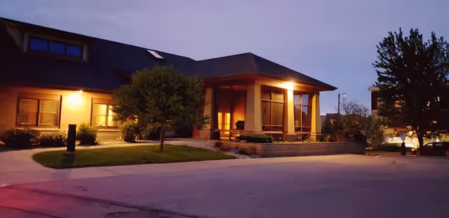 Exterior view of a single-story assisted living facility building at dusk with lights on inside and outside, a small tree and shrubs in front, and a paved driveway or parking area.