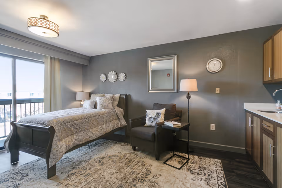 A cozy senior living bedroom featuring a single bed with patterned bedding, a dark armchair with a decorative pillow, a side table with books, a floor lamp, and a wall clock. The room has a large window with curtains allowing natural light, a decorative mirror on the wall, and wooden cabinetry with a countertop and sink on the right side. The floor has a patterned area rug.
