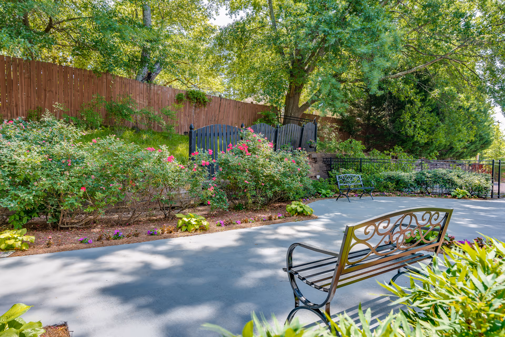 A peaceful outdoor garden area with a paved pathway, metal benches, flowering bushes, and tall trees providing shade. A wooden fence and a black gate are visible in the background.