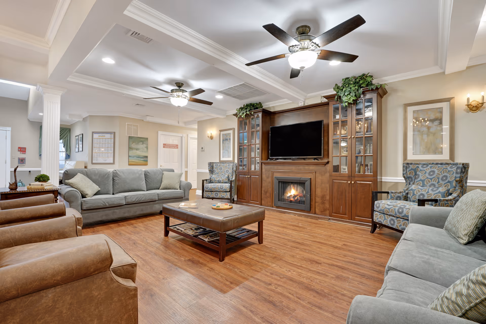 A spacious and well-lit living room with wooden flooring, two ceiling fans with lights, a large wooden entertainment center with a flat-screen TV and a fireplace, two gray sofas with cushions, two patterned armchairs, a brown leather ottoman coffee table, framed artwork on the walls, and decorative plants on top of the entertainment center.
