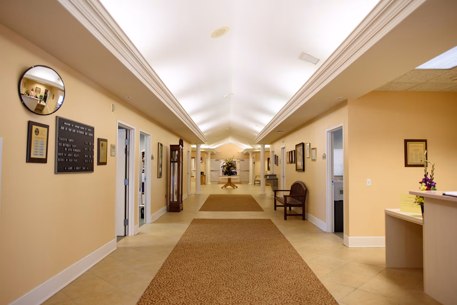 A long, well-lit hallway in a senior living facility with beige walls and tiled floors. The hallway features a patterned carpet runner, several doors on both sides, framed pictures and plaques on the walls, a wooden bench, a grandfather clock, and a round table with a flower arrangement at the far end. A reception desk with flowers is visible on the right side near the foreground.