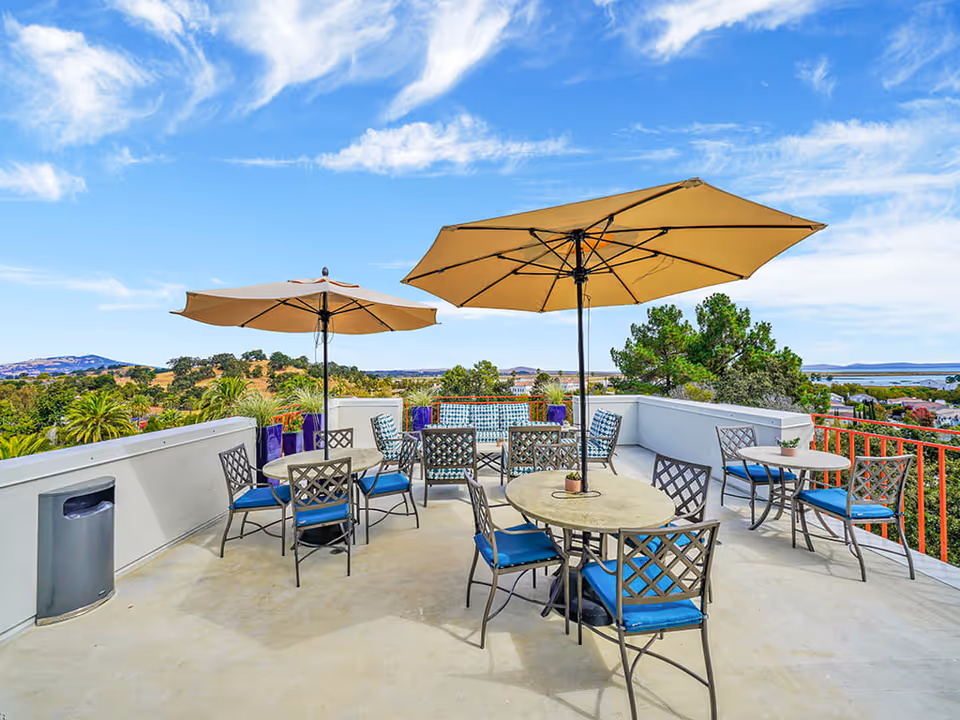 Outdoor patio area with several round tables and metal chairs with blue cushions. Two large beige umbrellas provide shade. The patio overlooks a scenic view of trees, hills, and a body of water under a blue sky with scattered clouds.