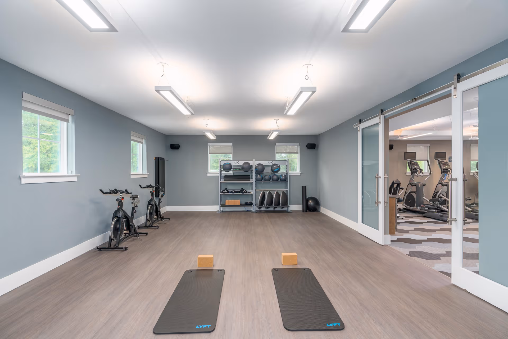 A fitness room with two black exercise mats and wooden blocks on a wooden floor, two stationary bikes along the left wall, and shelves with various exercise balls and equipment against the far wall. The room has light blue walls, three windows, and bright overhead lighting. Sliding glass doors on the right open to another room with cardio machines.