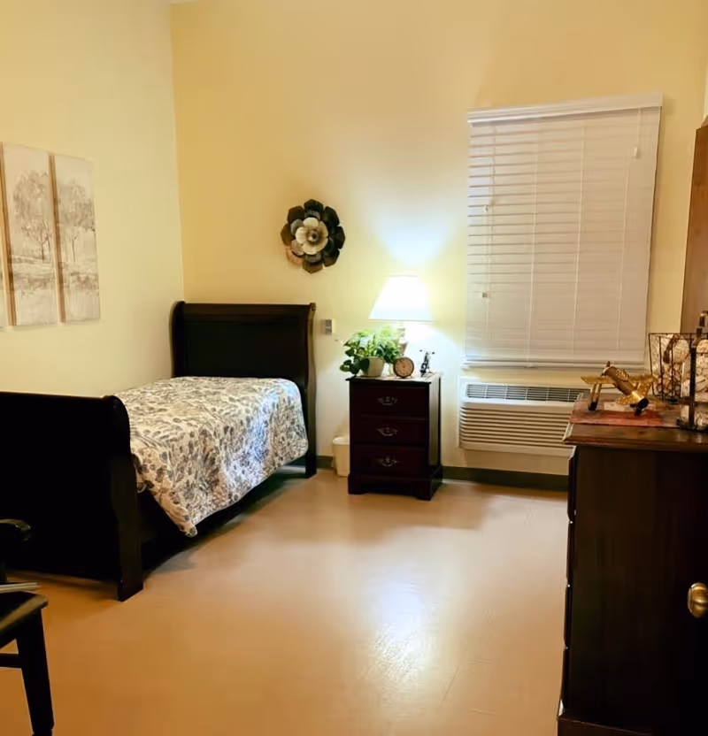 A cozy bedroom with a single dark wooden bed covered with a floral-patterned quilt. Next to the bed is a dark wooden nightstand with a lamp, a small clock, and a potted plant. A window with closed blinds and an air conditioning unit is on the wall behind the nightstand. On the right side, there is a dark wooden dresser with decorative items on top. The walls are light-colored, and there are two framed pictures and a flower-shaped wall decoration.