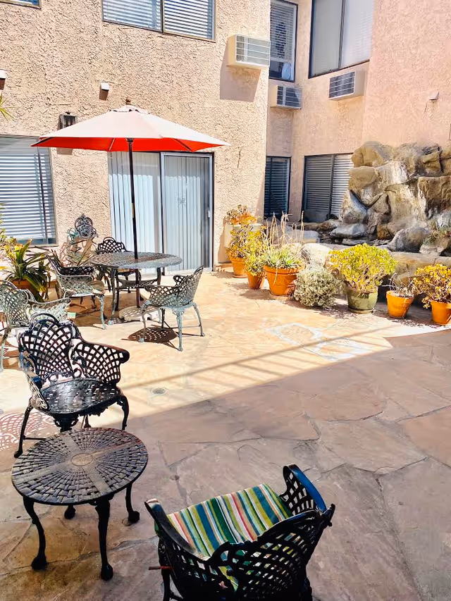 Outdoor patio area with metal chairs and tables, one table with a red umbrella. Several potted plants are arranged along the wall near a rock water feature. The patio is surrounded by beige building walls with windows and air conditioning units.