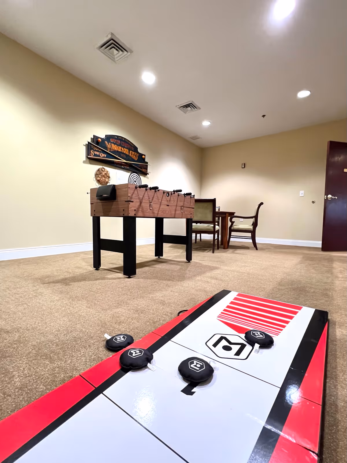 Indoor recreational room with a shuffleboard table in the foreground and a foosball table in the background. There is a small table with two chairs against the far wall, and a decorative wall sign above the foosball table. The room has beige walls, carpeted floor, and recessed ceiling lights.