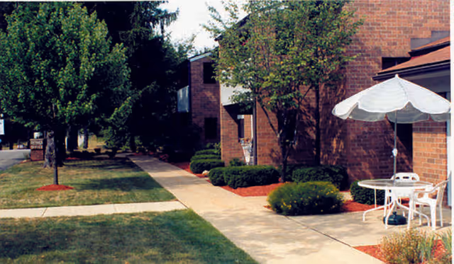 Outdoor view of a senior living facility with a brick building, a concrete walkway, green grass, trees, and a patio area with a white table, chairs, and an umbrella.
