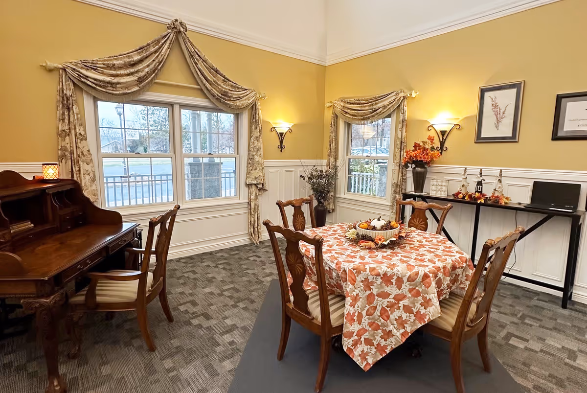 Cozy dining area with a round table covered in a fall-themed tablecloth, wooden chairs, a writing desk, windows with draped curtains, and seasonal decorations.
