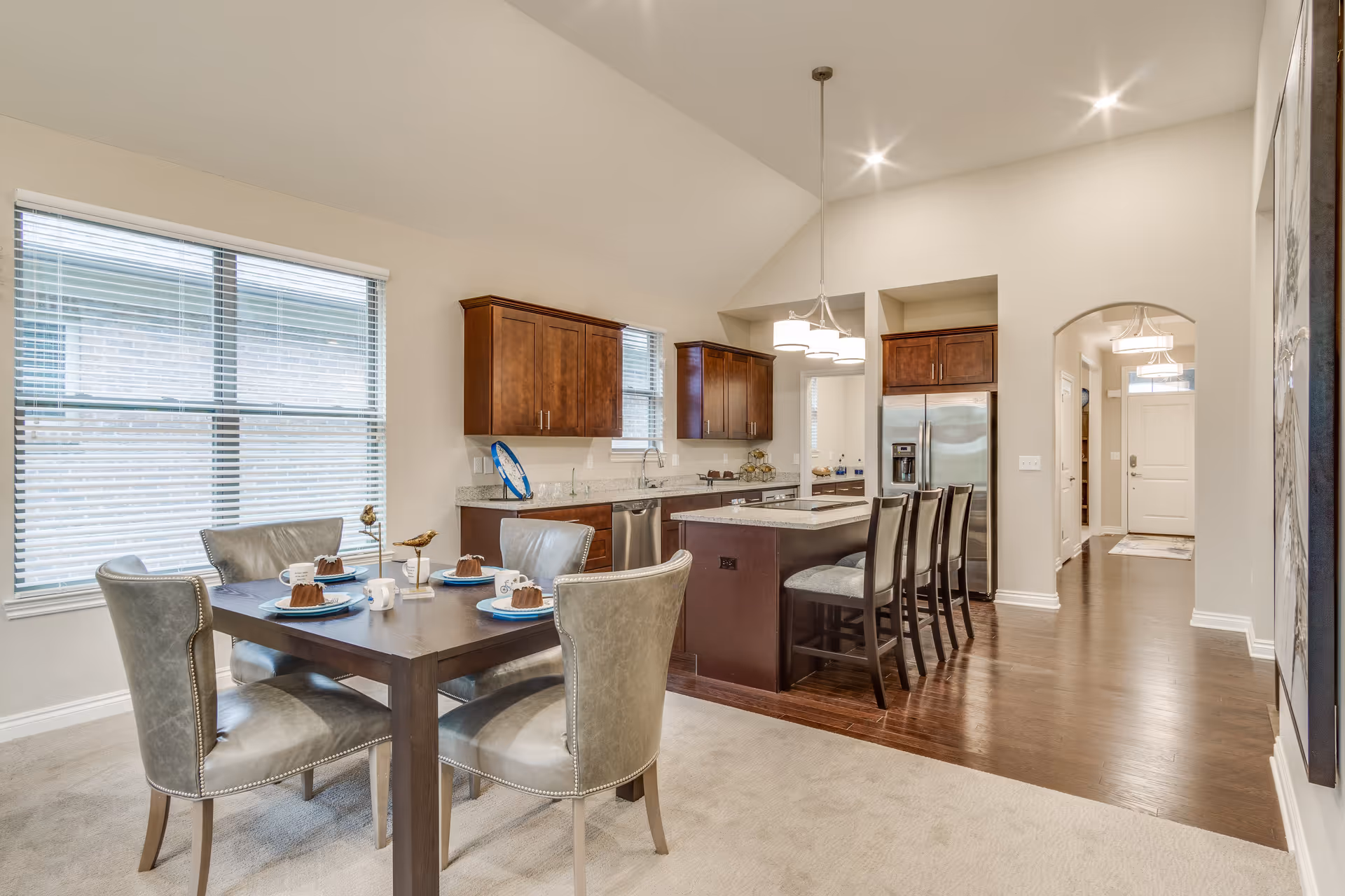 A bright and spacious kitchen and dining area in a senior living facility. The dining area features a dark wooden table set with four gray upholstered chairs and place settings including plates, mugs, and small cakes. The kitchen has dark wooden cabinets, a large island with three bar stools, stainless steel appliances including a refrigerator and dishwasher, and granite countertops. Large windows with blinds allow natural light to fill the room, and recessed lighting and a hanging light fixture illuminate the space. The flooring transitions from carpet in the dining area to wood in the kitchen and hallway leading to the front door.