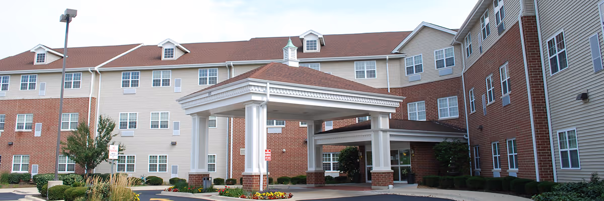 Exterior view of a multi-story senior living facility with a covered entrance, brick and siding facade, multiple windows, and landscaped greenery around the driveway.