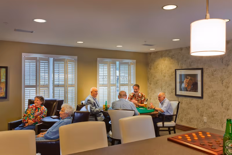 Several elderly residents sit in a bright lounge area around a table playing cards while others relax in armchairs.