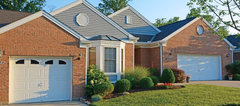Front exterior of a brick residential building with two white garage doors, a bay window, and neatly landscaped lawn and shrubs.
