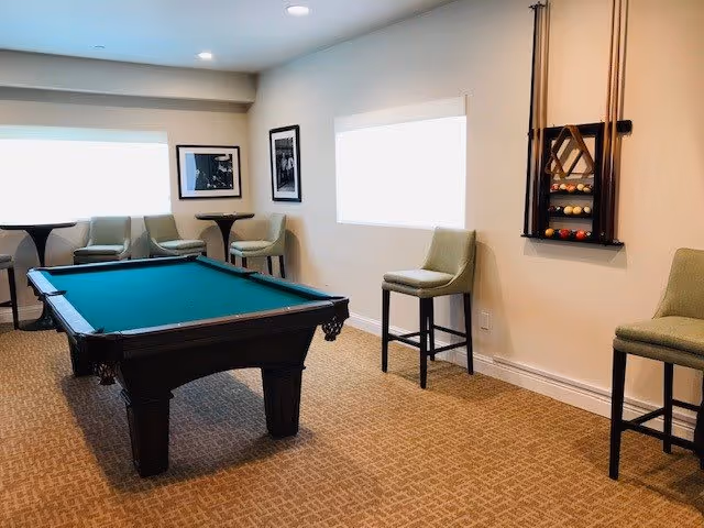 A recreational room with a pool table in the center, surrounded by several green cushioned chairs and small round tables. The walls are light-colored with two framed black and white photographs and a wall-mounted pool cue rack holding cues and billiard balls. The room is carpeted and well-lit with recessed ceiling lights.