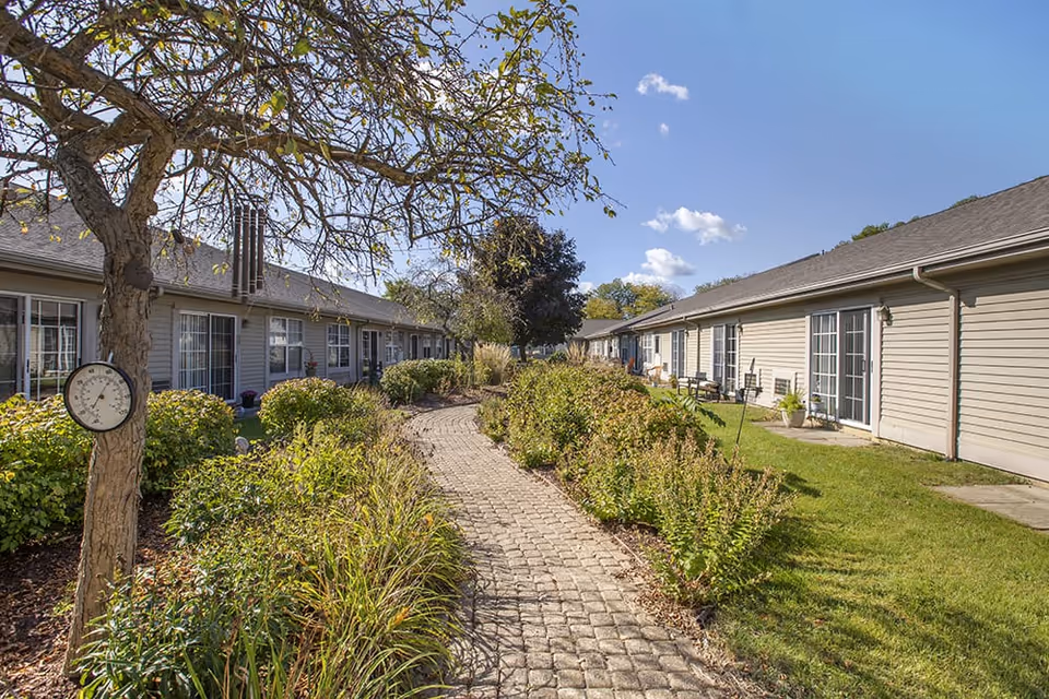 A cobblestone path winds through a landscaped courtyard between single-story residential wings with patio doors under a blue sky.