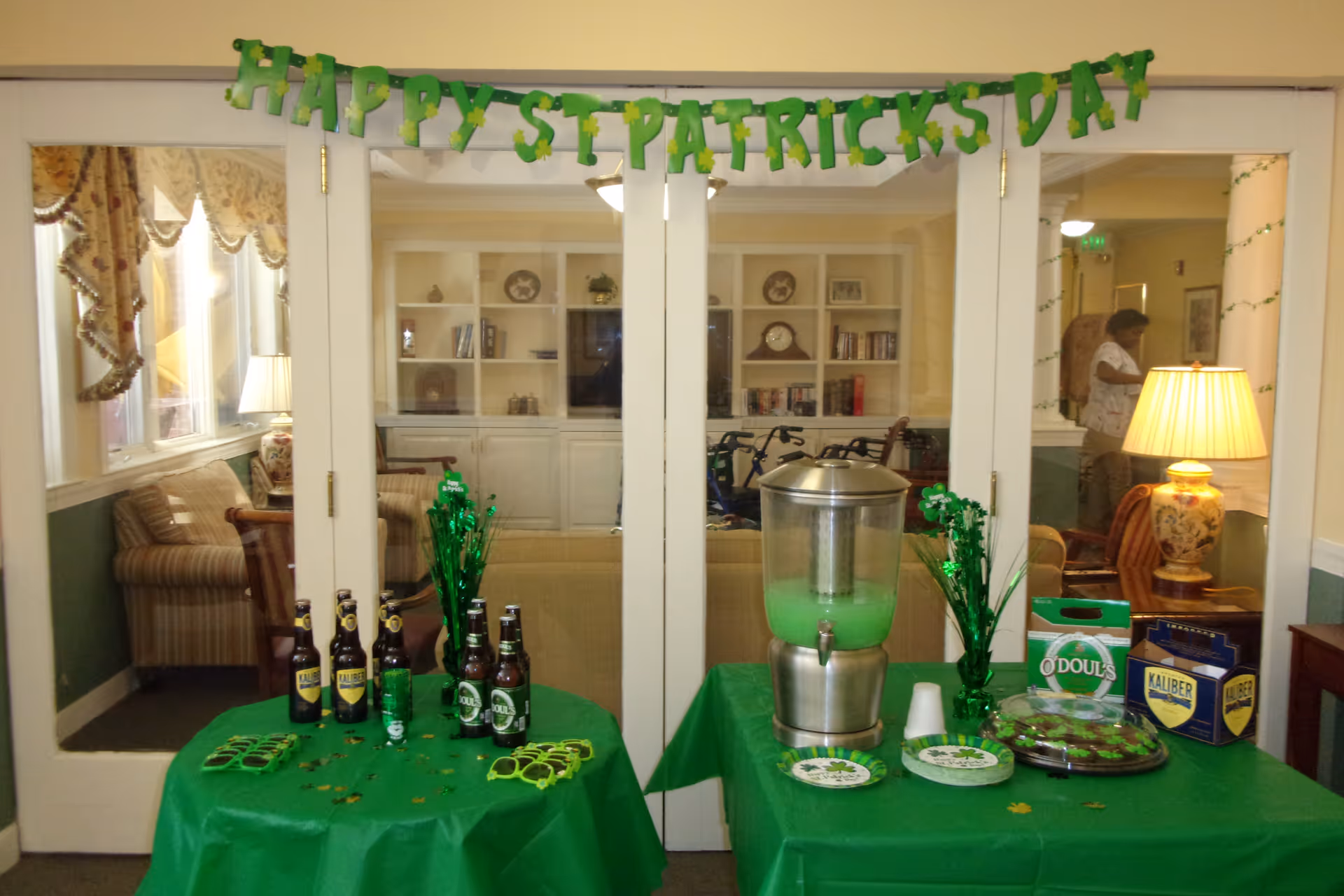 A festive St. Patrick's Day setup in a senior living facility with two tables covered in green tablecloths. One table has bottles of non-alcoholic beer, green shamrock decorations, and green sunglasses. The other table has a large beverage dispenser with green liquid, plates, cups, a box of O'Doul's non-alcoholic beer, a box of Kaliber beer, and a tray of decorated cookies. Behind the tables are glass doors leading to a cozy sitting area with armchairs, lamps, and bookshelves. A green banner above the doors reads 'HAPPY ST PATRICKS DAY'.