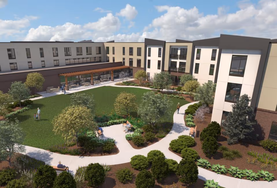 Outdoor courtyard area of a senior living facility with a large green lawn, walking paths, benches, trees, and shrubs. The building surrounds the courtyard with multiple windows and a covered patio area with seating. Several people are seen walking or sitting in the courtyard.