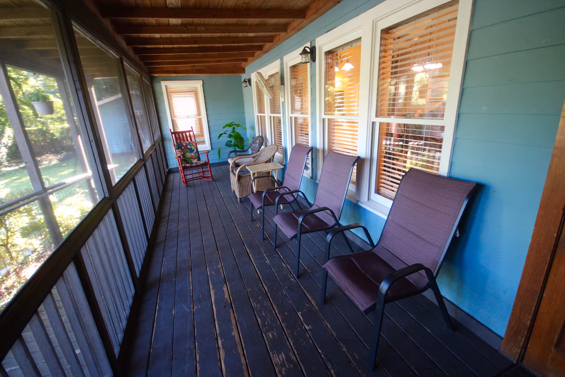 A screened-in porch with wooden floorboards and ceiling beams. The porch has four purple cushioned chairs lined up against a blue wall with three windows. At the far end, there is a red rocking chair with a floral cushion and a wicker chair with a small table. A green potted plant is placed near the window at the end of the porch.