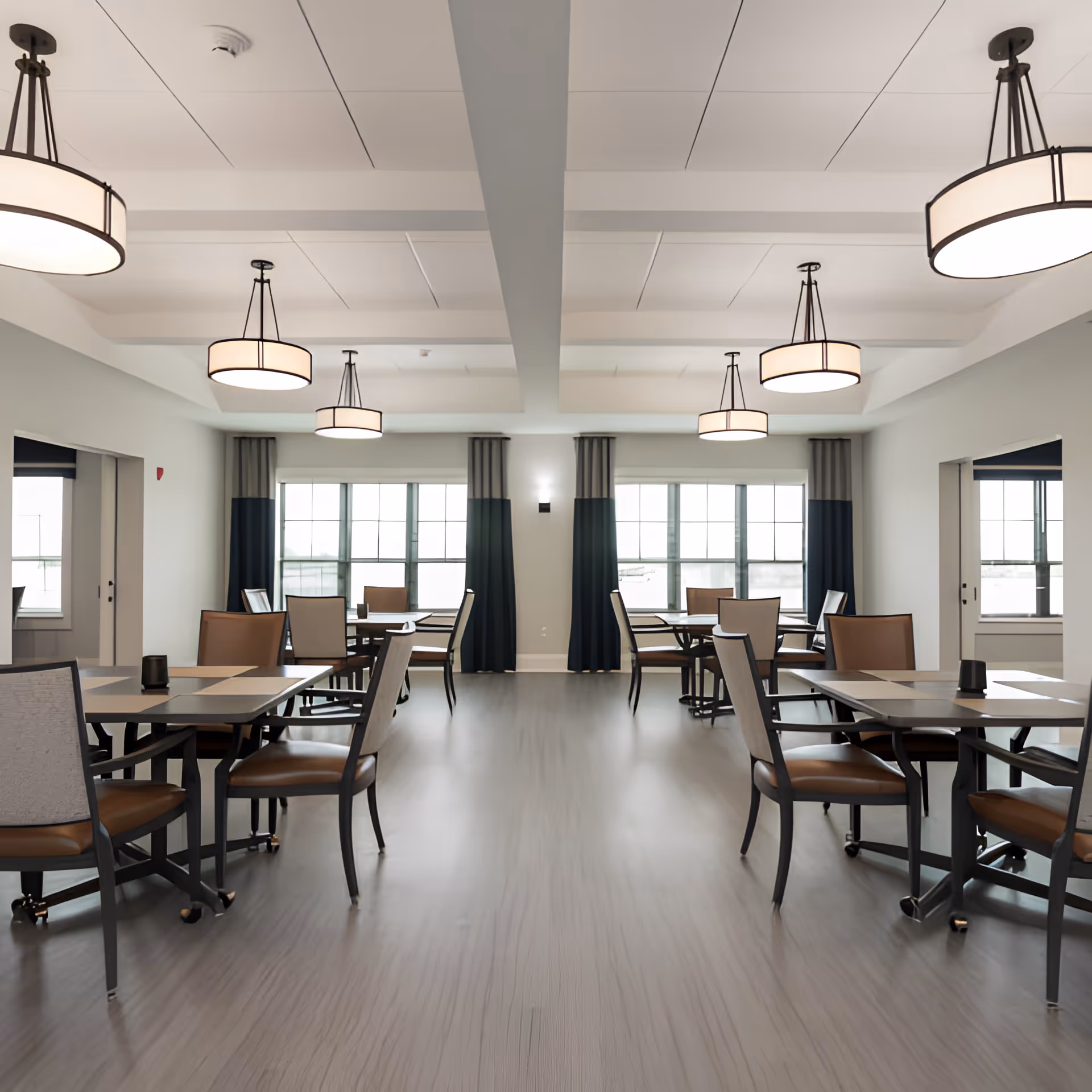 A bright and spacious dining room in a senior living facility with multiple tables and chairs arranged neatly. The room features large windows with two-tone curtains allowing natural light to fill the space. Modern ceiling lights hang from a white ceiling with beams, and the floor has a light wood finish.