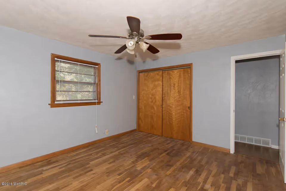 Empty bedroom with hardwood floors, a ceiling fan, a window with blinds, and a wooden closet.